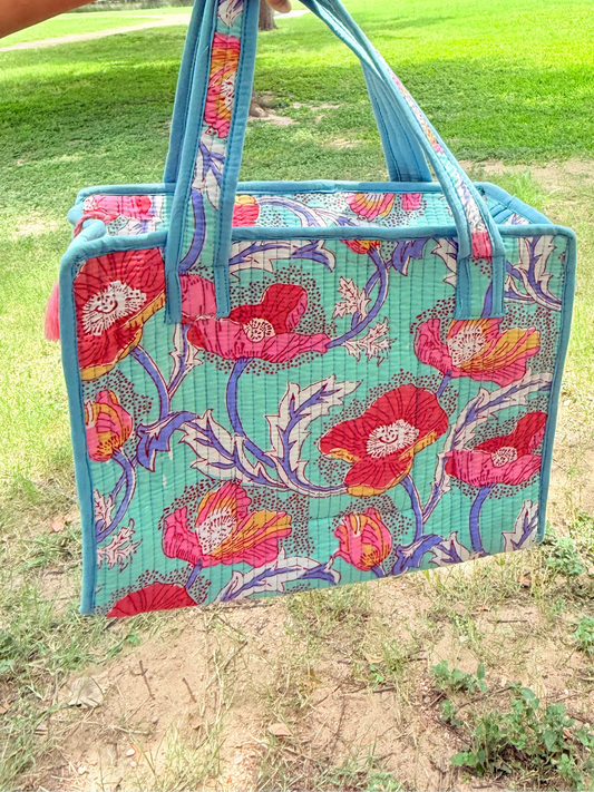 A woven tote bag with a turquoise blue base and a vibrant red, pink, and purple floral pattern, held by a person's arm in an outdoor park setting.